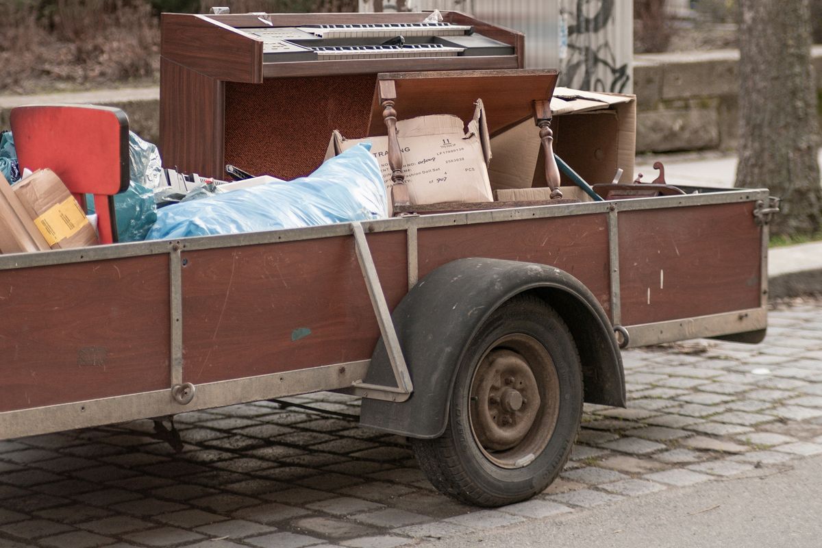 Aanhangwagen op een kasseistraat, geladen met een oud orgel, een rode stoel, dozen en zakken