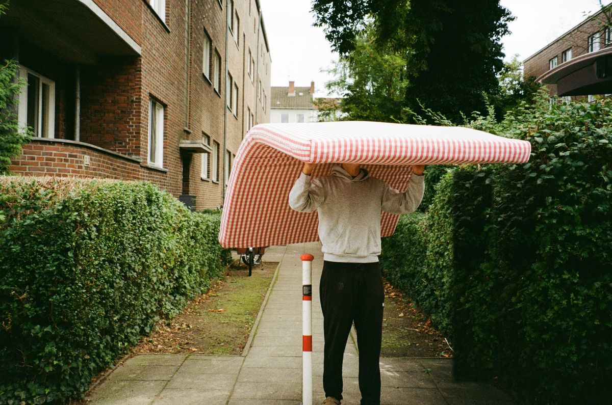 Iemand draagt een matras boven het hoofd over een stoep tussen twee hagen